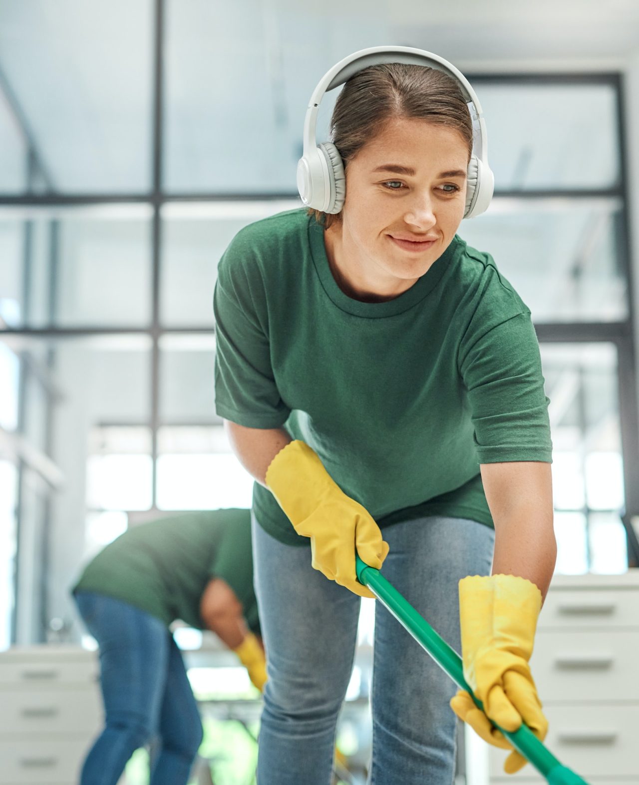 shot of a young woman cleaning an office with her colleague in the background